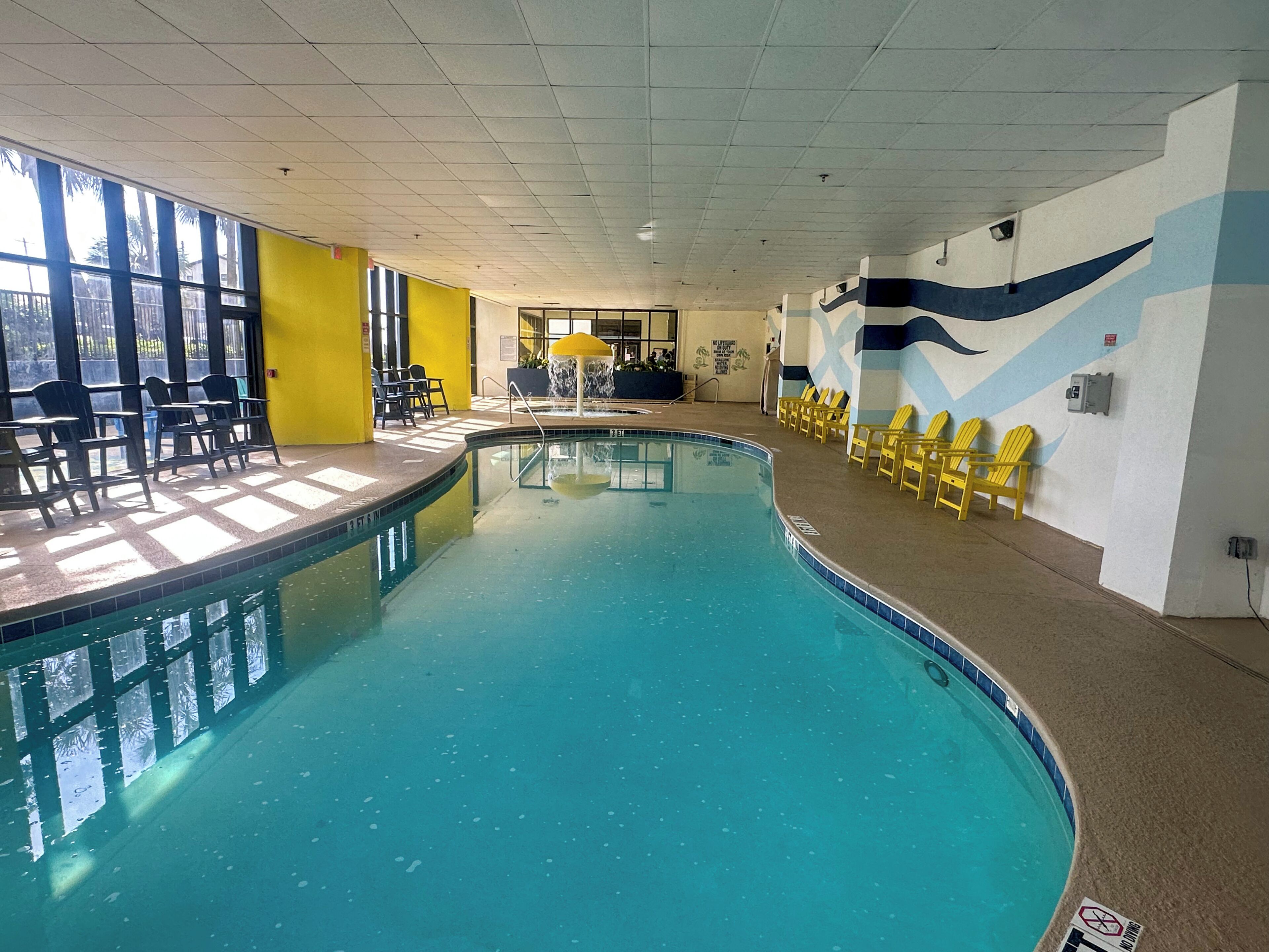 Une piscine intérieure avec des chaises jaunes, une fontaine et une vue sur des palmiers à travers de grandes fenêtres.