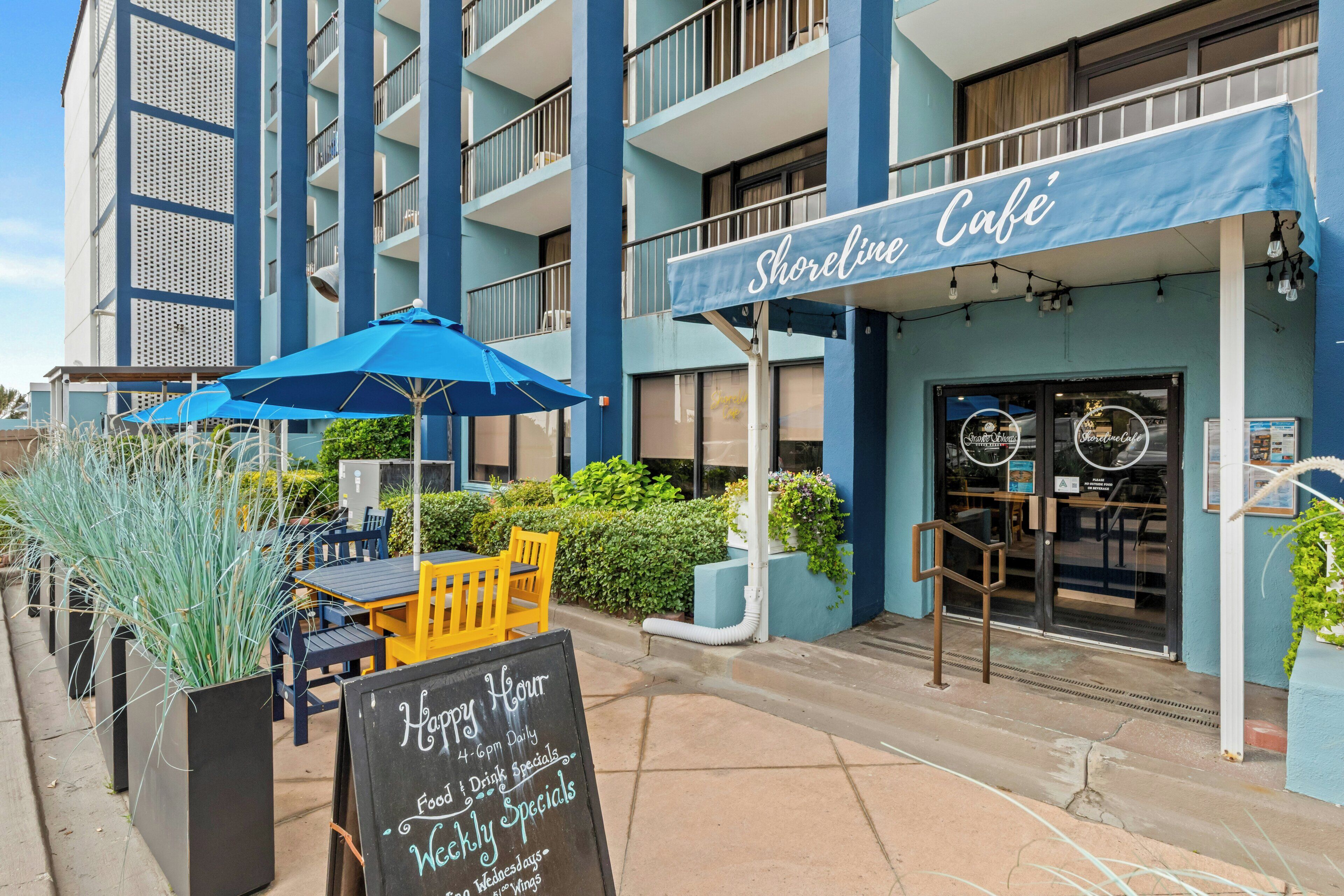 A beachfront café with outdoor seating, a chalkboard sign advertising specials, and a blue awning.