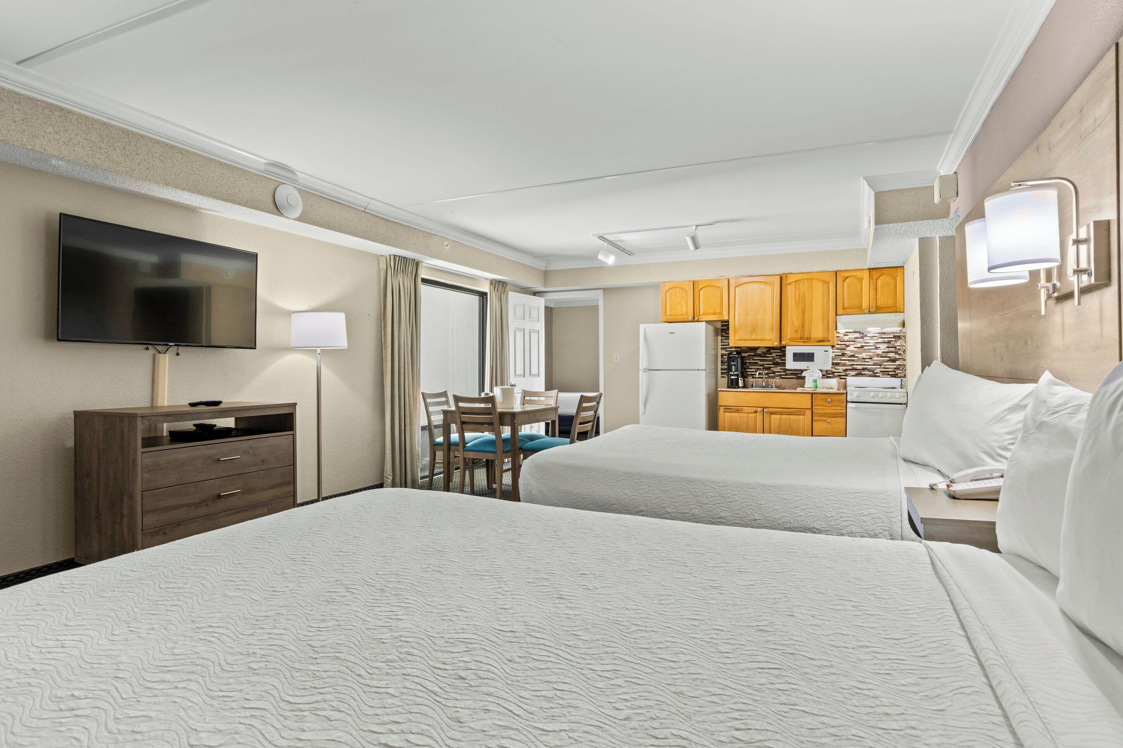 A hotel room with two beds, a TV, a desk with a chair, a kitchenette, and a bathroom.