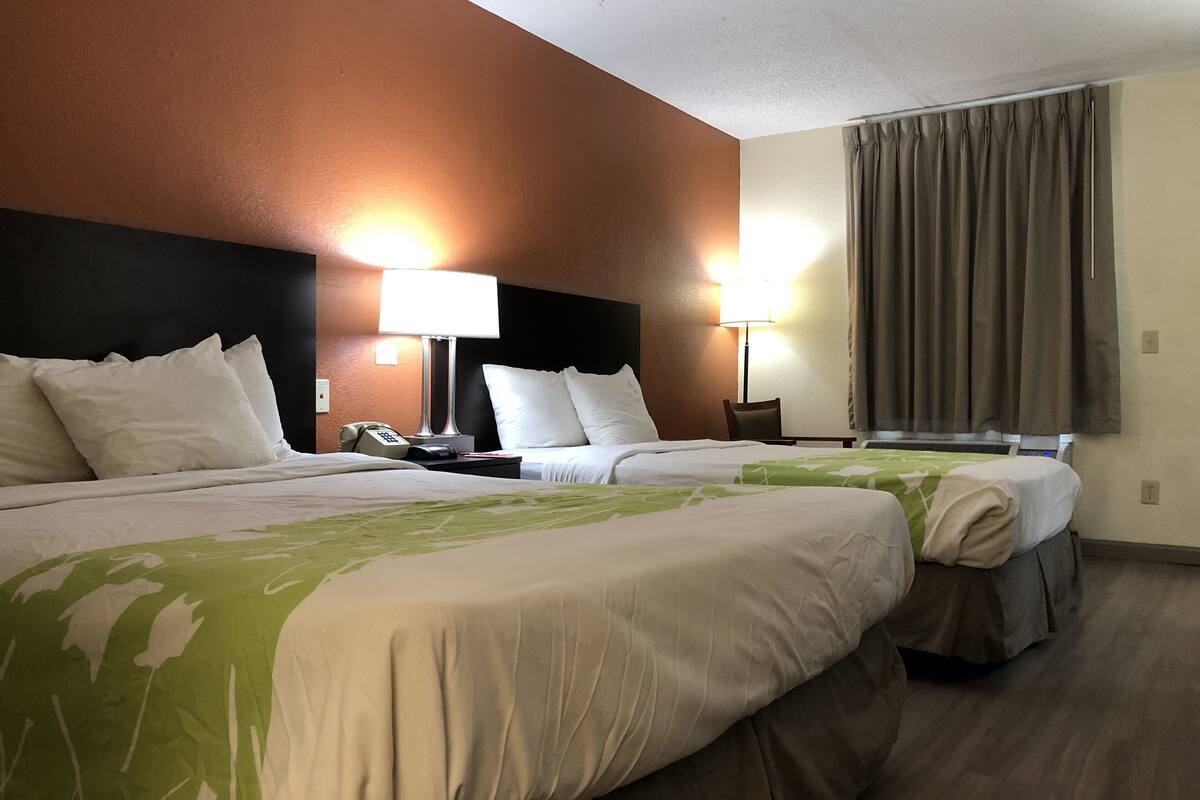 Habitación de hotel con dos camas, una mesita de noche con lámpara, una silla y una ventana con cortinas.