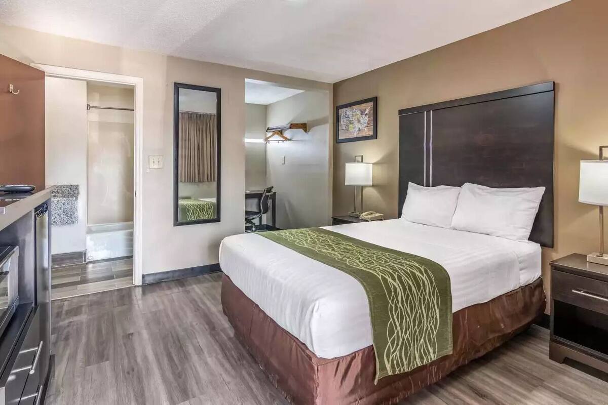A hotel room with a large bed, a desk, a chair, a mirror, a bedside table with a lamp, and a wardrobe.