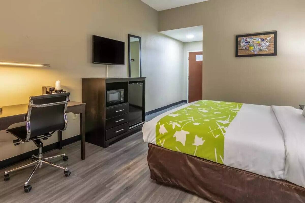 A hotel room with a bed, a desk with a chair, a television, a mirror, and a framed picture on the wall.