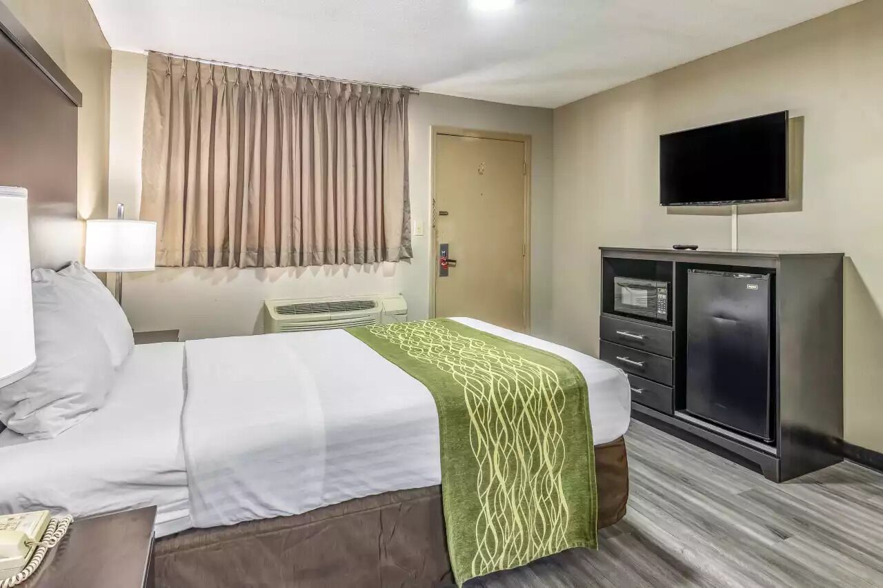 A hotel room with a bed, a desk with a lamp, a TV mounted on the wall, and a minibar.