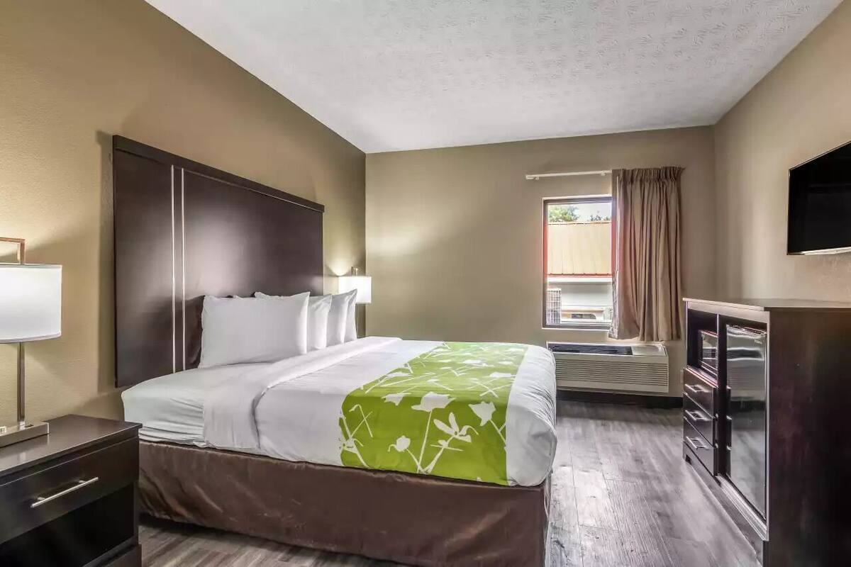 A hotel room with a bed, bedside table, lamp, air conditioning unit, and a TV.