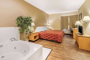 Habitación de hotel con una bañera grande, una cama con colcha roja, un escritorio con ordenador y una planta en maceta.