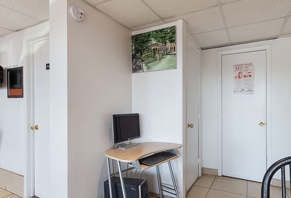 Una pequeña oficina con un escritorio, computadora y silla. Hay una puerta con un cartel, una cámara de seguridad instalada en la pared y un póster.