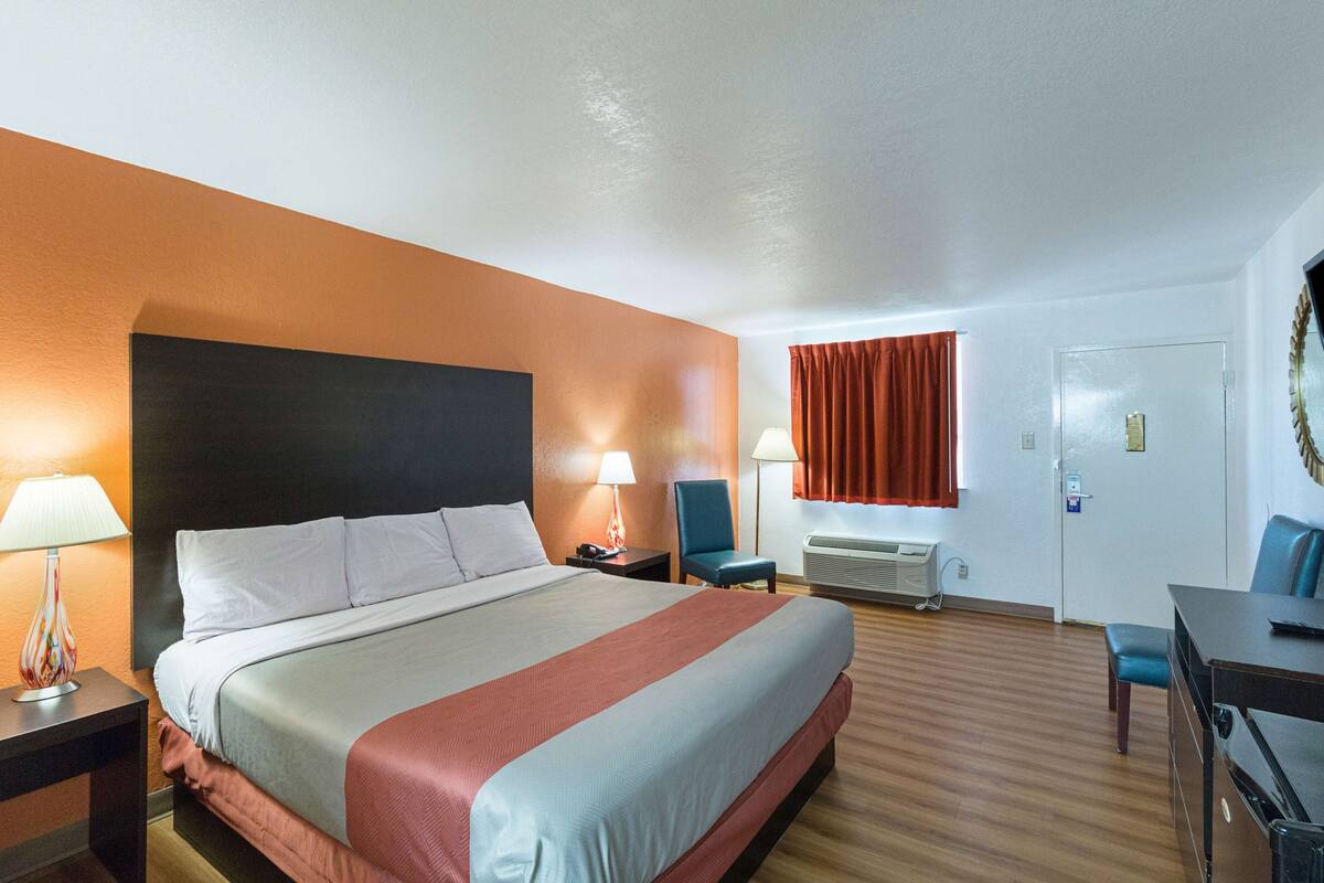 A hotel room with a large bed, two bedside tables with lamps, a desk with a chair, an air conditioning unit, and a door with a mirror.