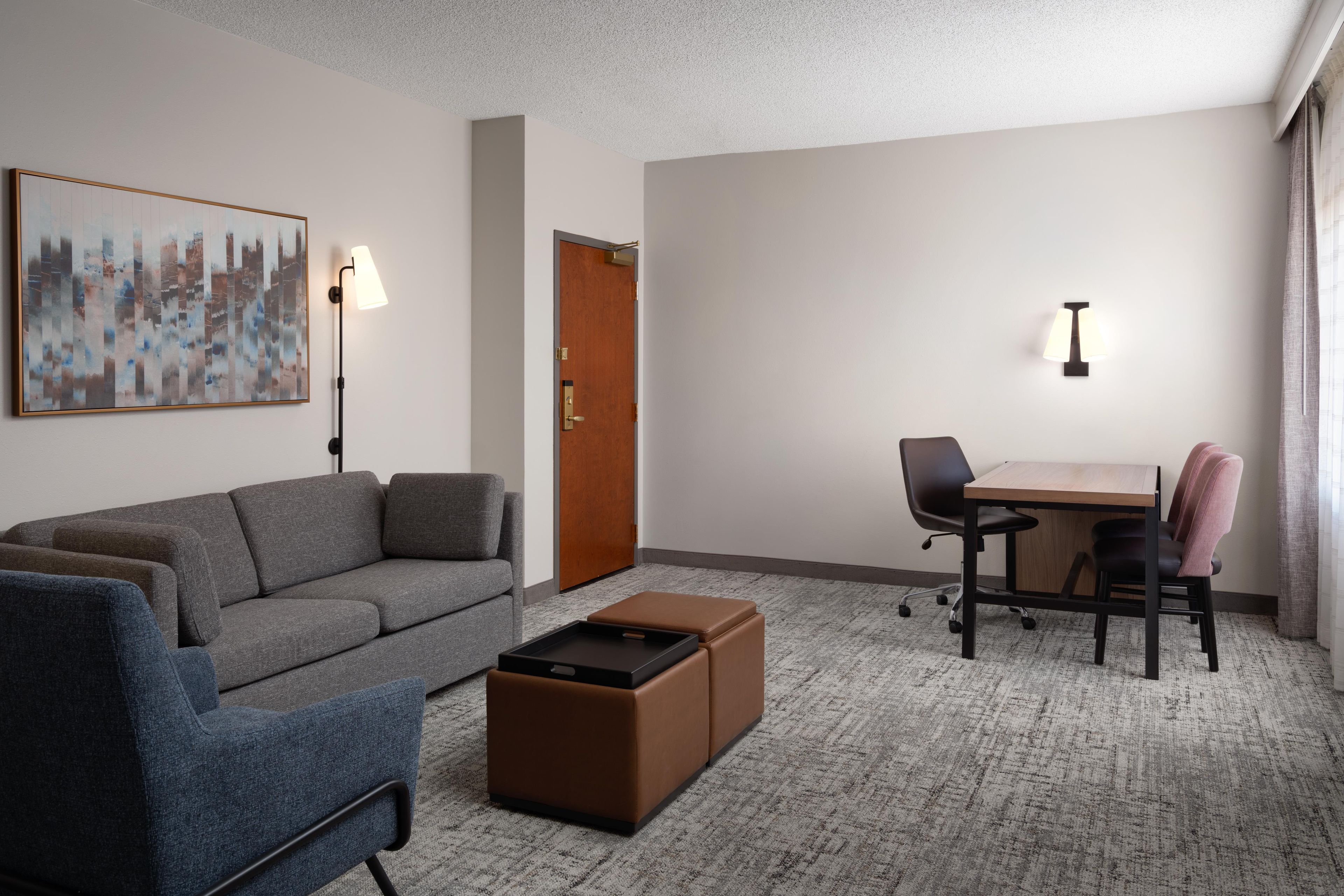 A hotel room with a grey sofa, a small brown ottoman, a desk with two chairs, a wall-mounted lamp, and a door.
