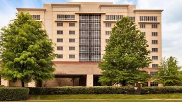 A multi-story hotel with a central glass facade, surrounded by well-maintained hedges and trees.