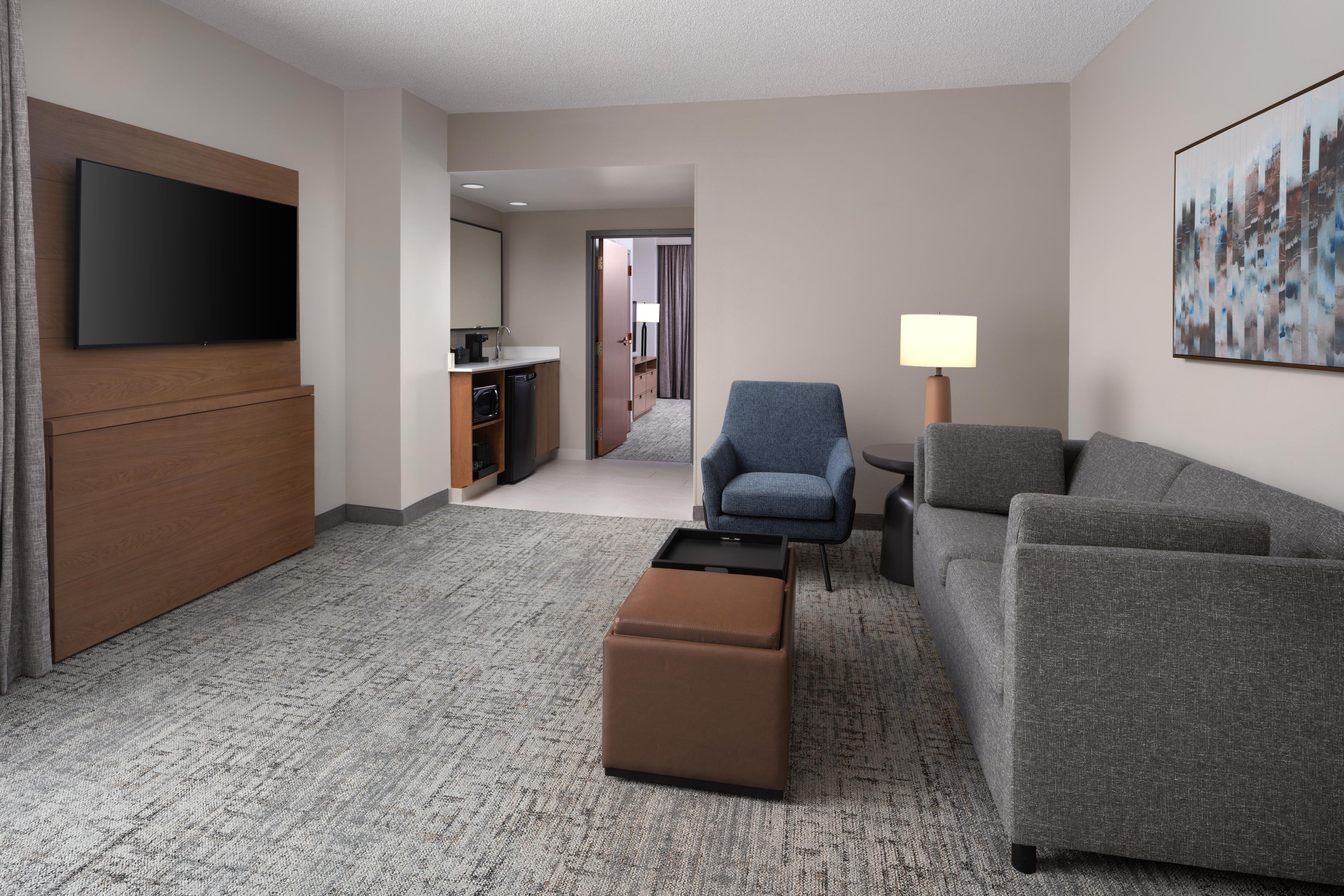 A hotel room with a flat-screen TV, a sofa, a chair, a small ottoman, a lamp, and a painting on the wall.