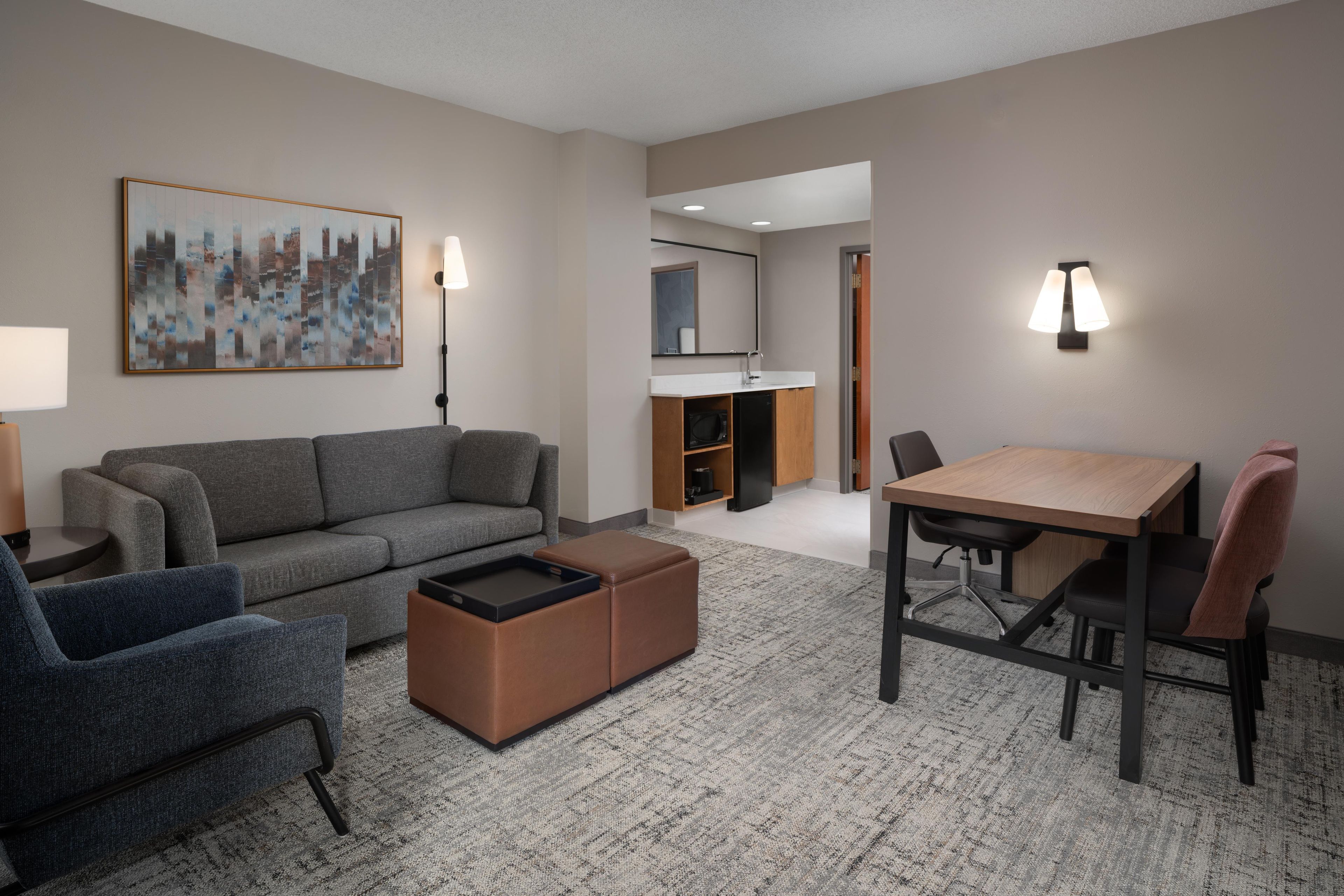A modern hotel room with a grey sofa, a small dining table, a blue armchair, and a brown ottoman.