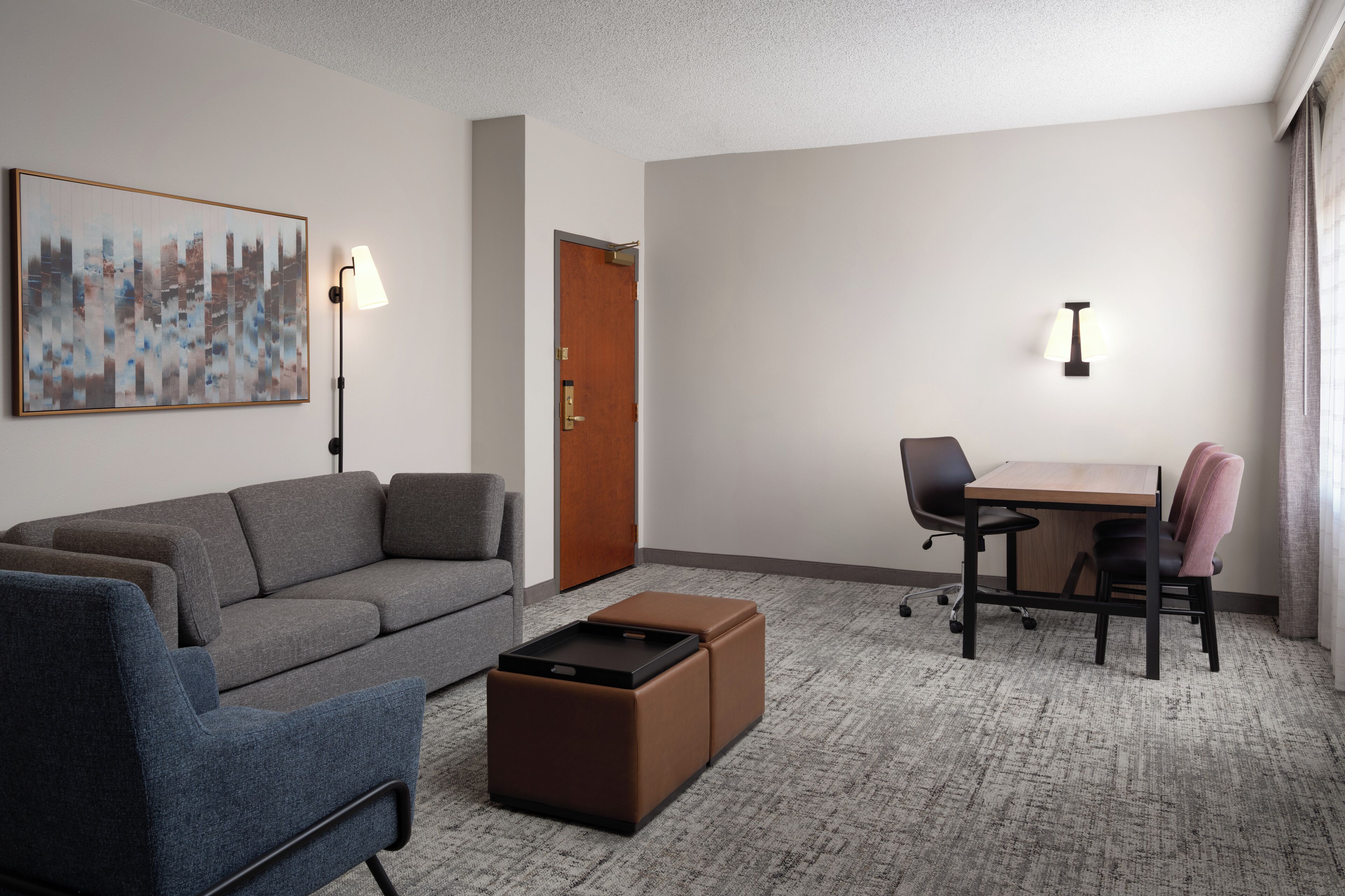 A hotel room with a gray sofa, a small ottoman, a wooden table with two chairs, a lamp, and a door.
