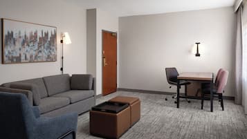 A hotel room with a gray sofa, a small ottoman, a wooden table with two chairs, a lamp, and a door.
