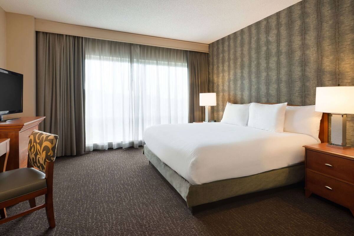 A hotel room with a large bed, a TV, a desk with a chair, and a nightstand with a lamp.