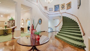 Un amplio vestíbulo con una imponente escalera, una mesa redonda con jarrones y un candelabro.
