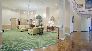Un vestíbulo espacioso con una alfombra de diseño verde, un sofá, una mesa con un jarrón, un reloj y una escalera que conduce a un nivel superior.