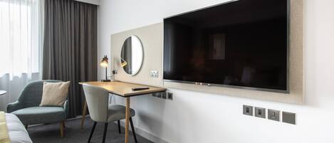 Une chambre d’hôtel comprenant un lit, un bureau avec une chaise, une grande télévision, un miroir et une fenêtre avec des rideaux.