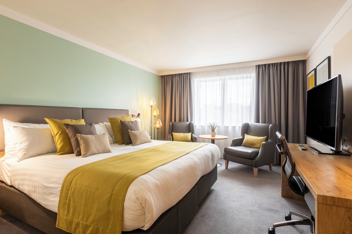 Une chambre d’hôtel avec un grand lit, deux fauteuils, un bureau avec une télévision et une petite table avec une lampe.