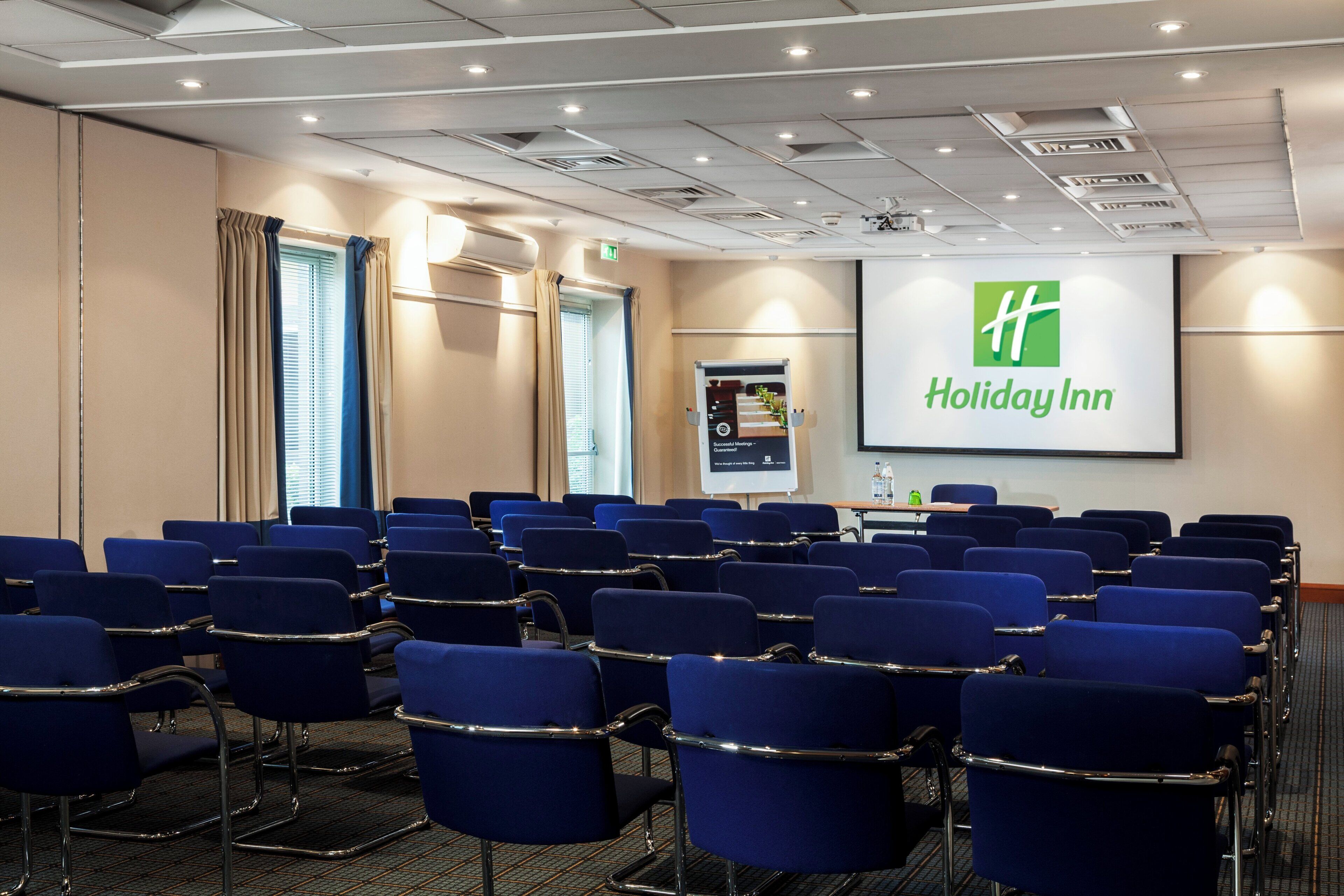Une salle de conférence avec des rangées de chaises bleues, un écran de projection orné du logo de Holiday Inn et un écran de présentation.
