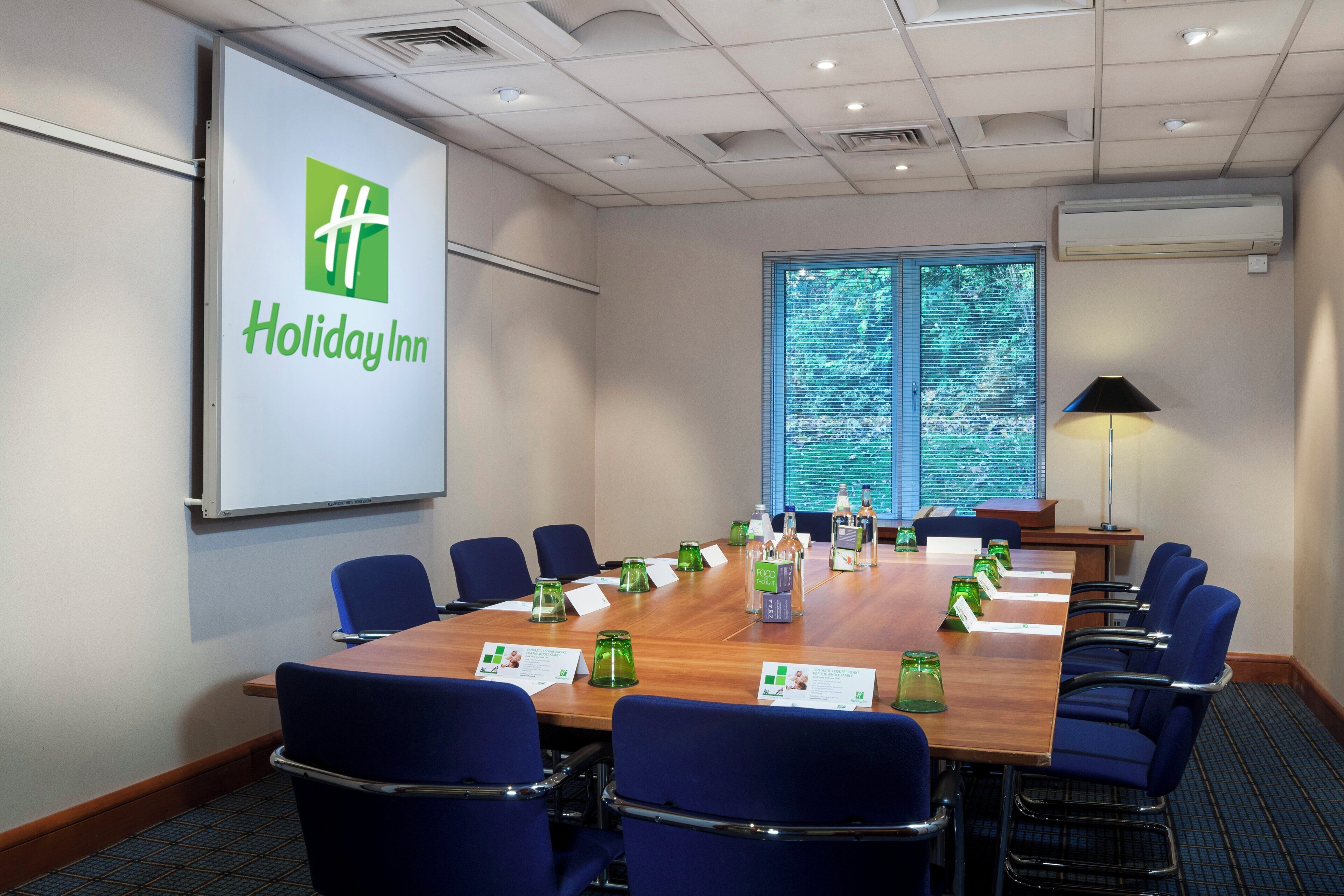 A conference room with a long table, blue chairs, a projector screen with the Holiday Inn logo, and a large window with blinds.