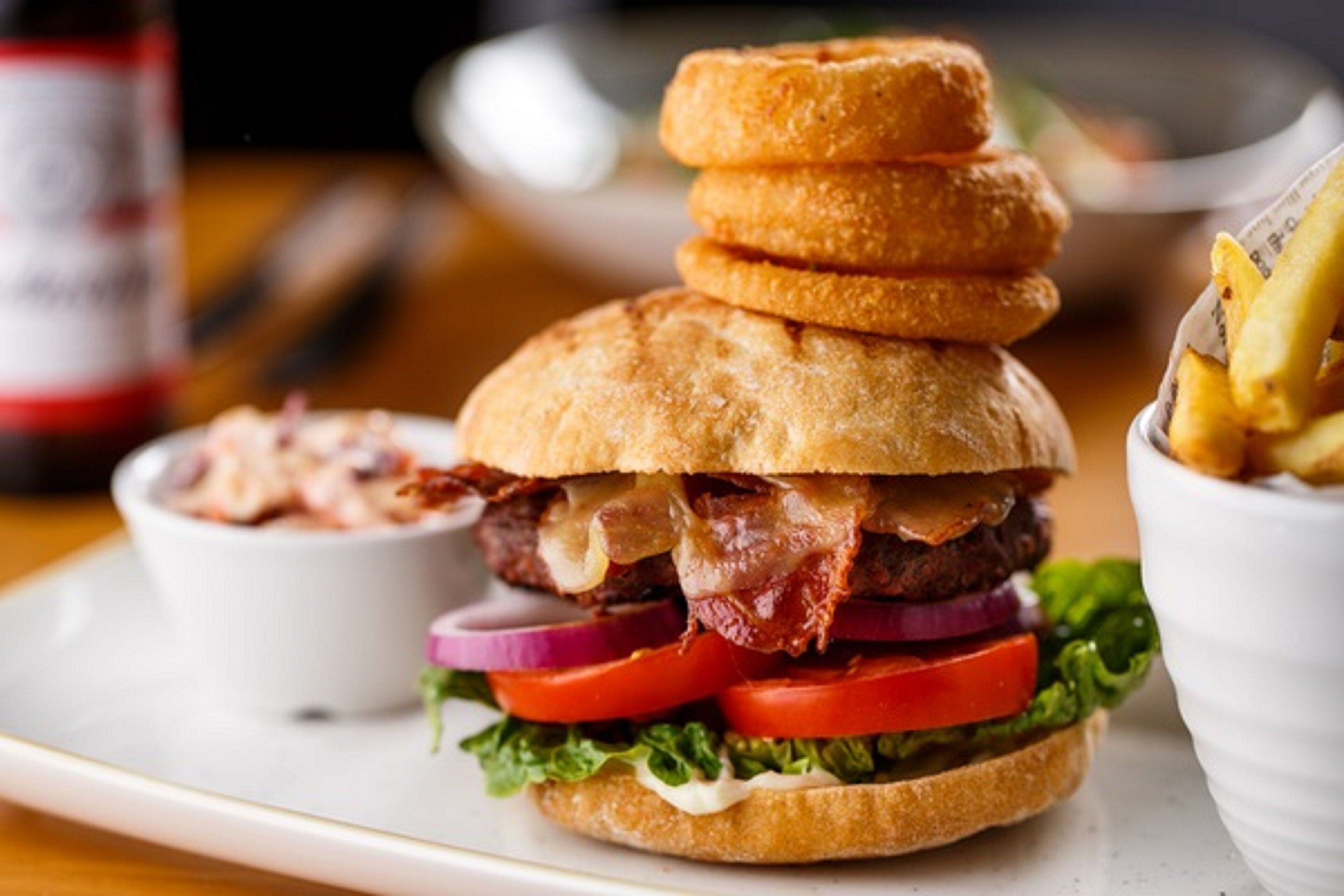 A burger with stacked buns, lettuce, tomato, red onion, and bacon, served with a side of fries and a small bowl of coleslaw.