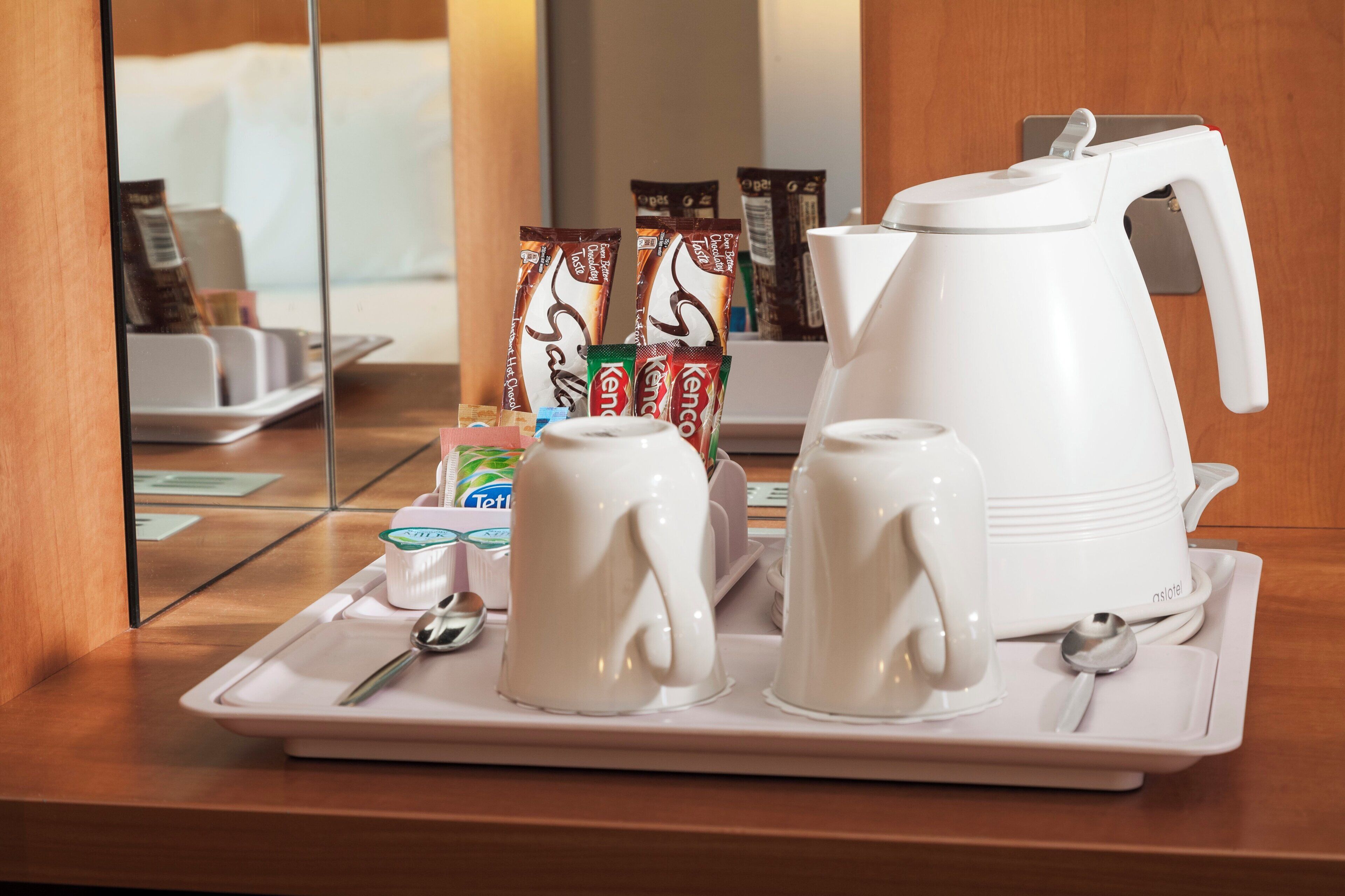 A tray with a white teapot, two white cups, a spoon, and several packets of instant coffee on a wooden surface.
