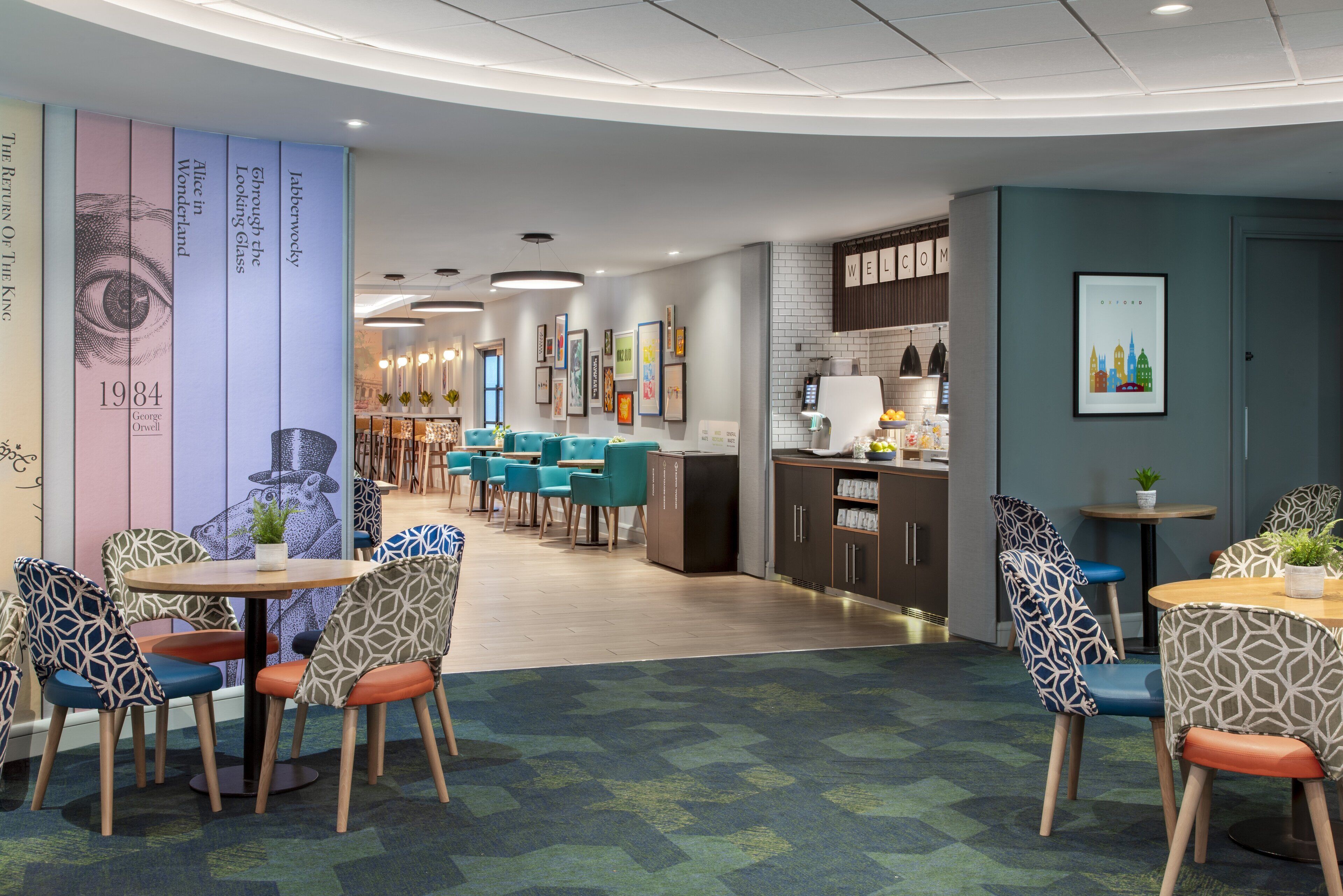 A modern dining area with patterned chairs and tables, a kitchen area with a refrigerator and cabinets, and a wall with framed pictures and a 'WELCOME' sign.