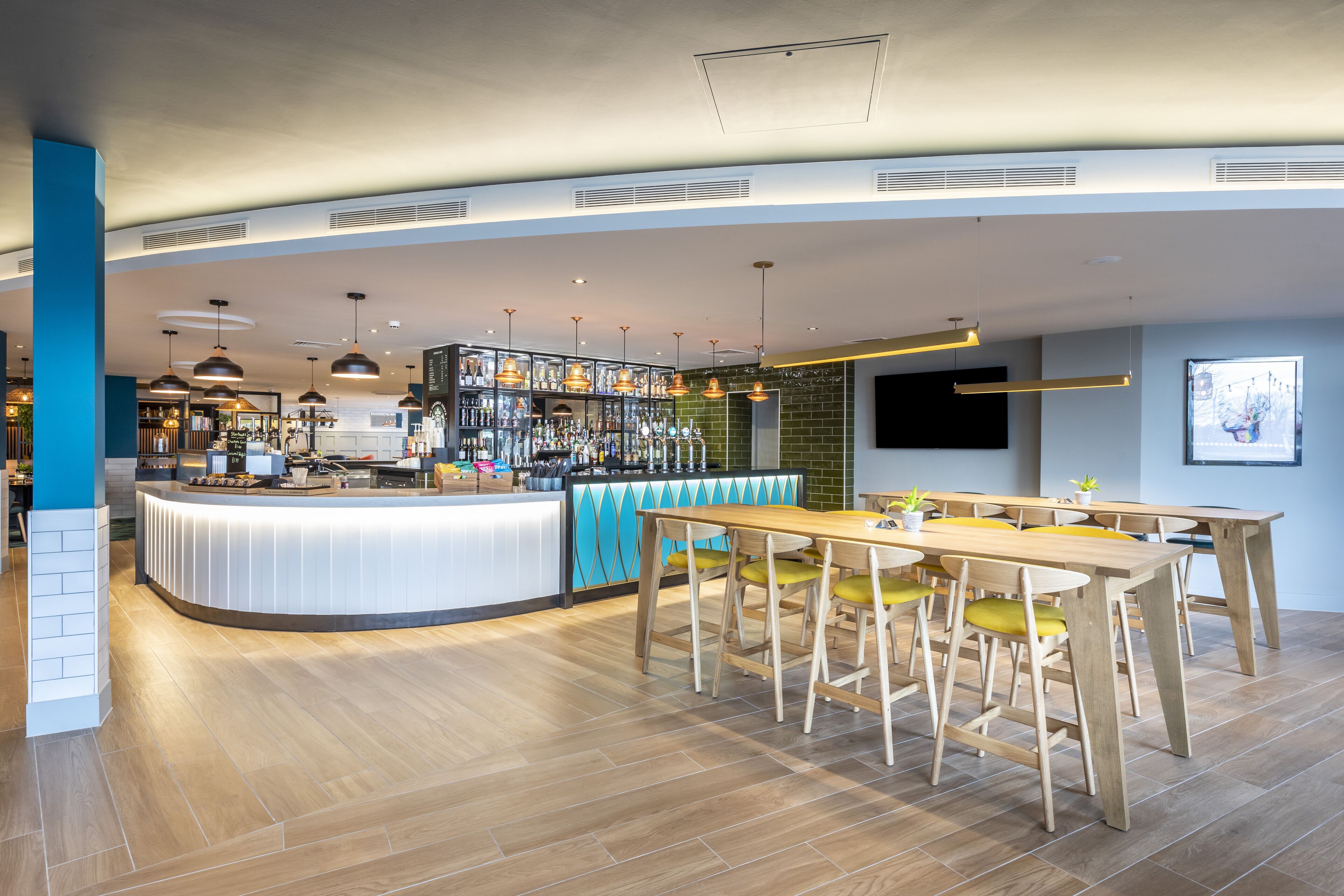 A modern bar area with a curved counter, wooden tables, and yellow chairs.