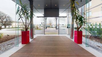 Una entrada de edificio moderna con una rampa de madera que conduce a una puerta de vidrio, rodeada de macetas con plantas y una vista despejada de la calle.