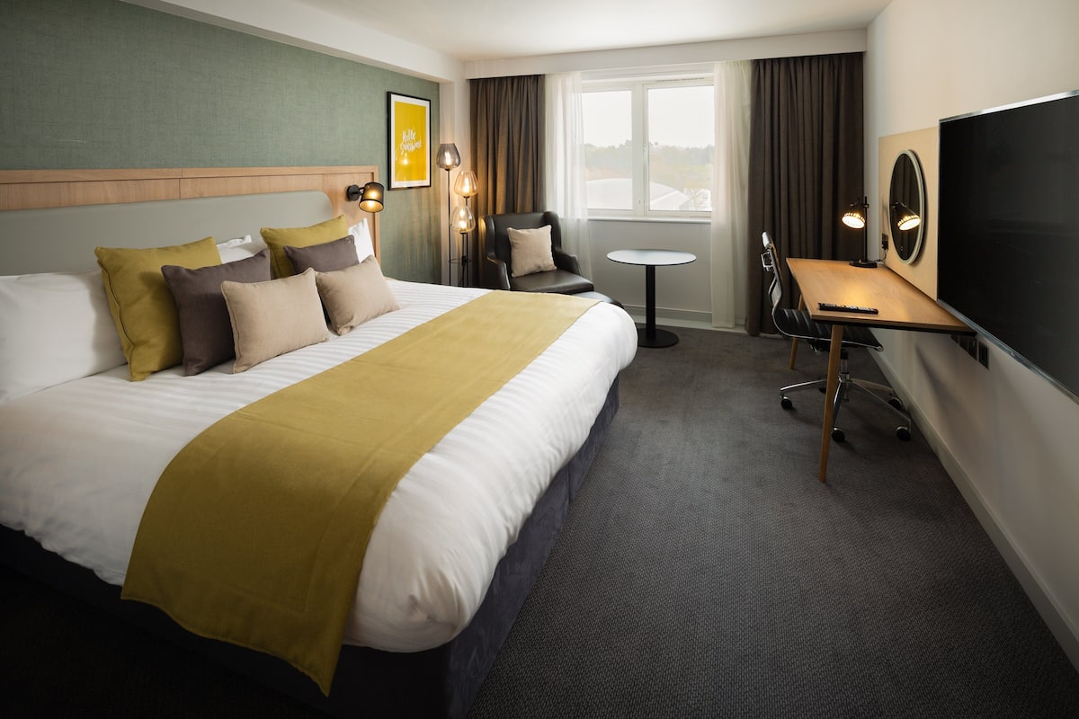 A hotel room with a large bed, a desk, a chair, a TV, and a window with curtains.