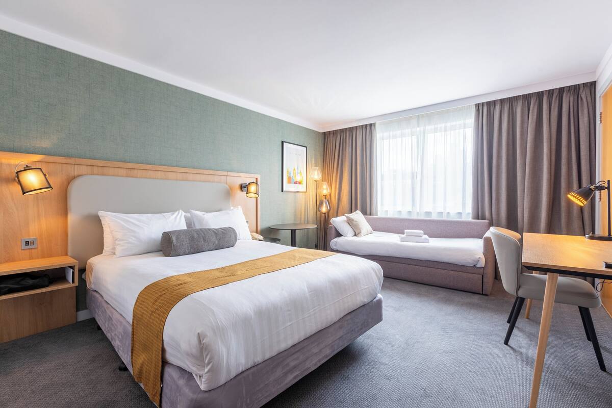 A hotel room with a large bed, a smaller bed, a desk, and a chair. There is a window with curtains, a lamp, and a picture on the wall.