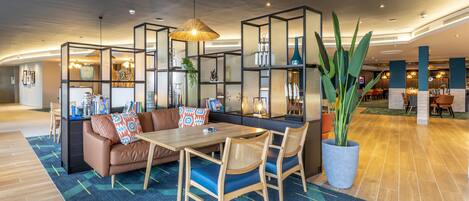 A modern hotel lobby with a seating area featuring a wooden table and chairs, a sofa with patterned cushions, and a large potted plant.