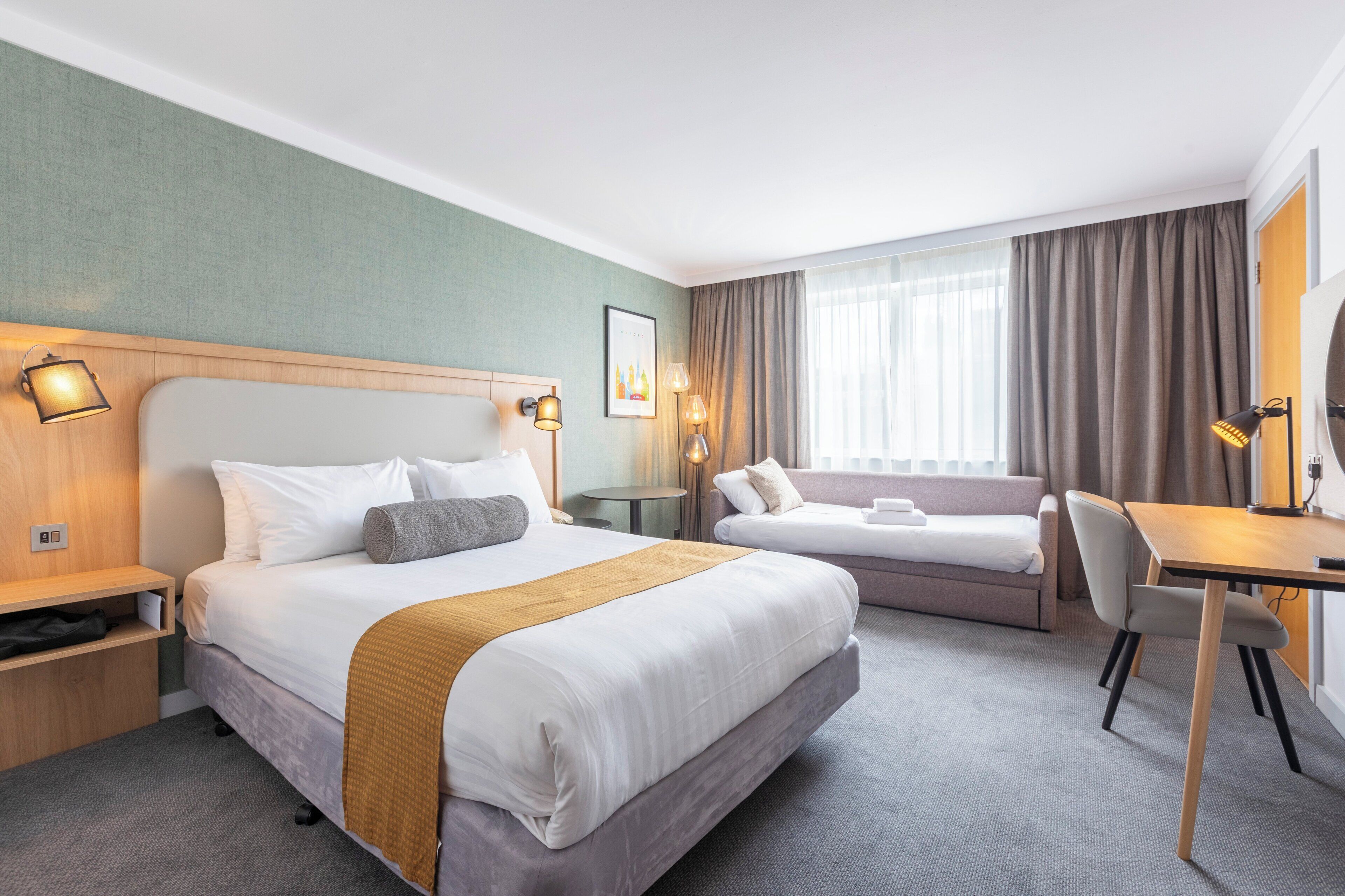 A hotel room with a large bed, a smaller bed, a desk, and a chair. There are two bedside tables with lamps.