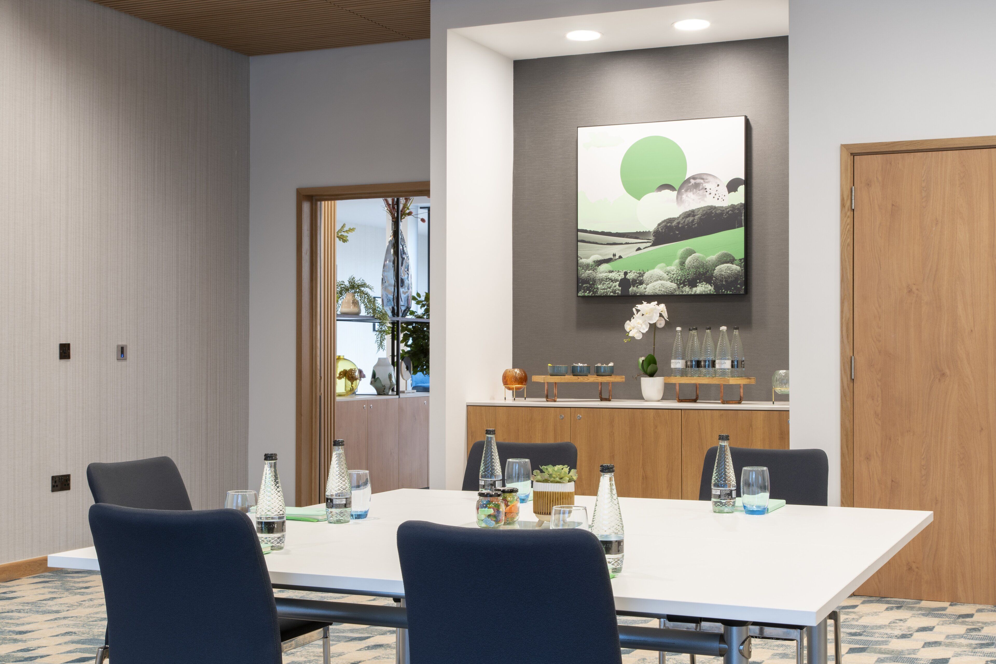 Un salón de conferencias con una larga mesa, sillas, un cuadro y un aparador de madera con objetos decorativos.