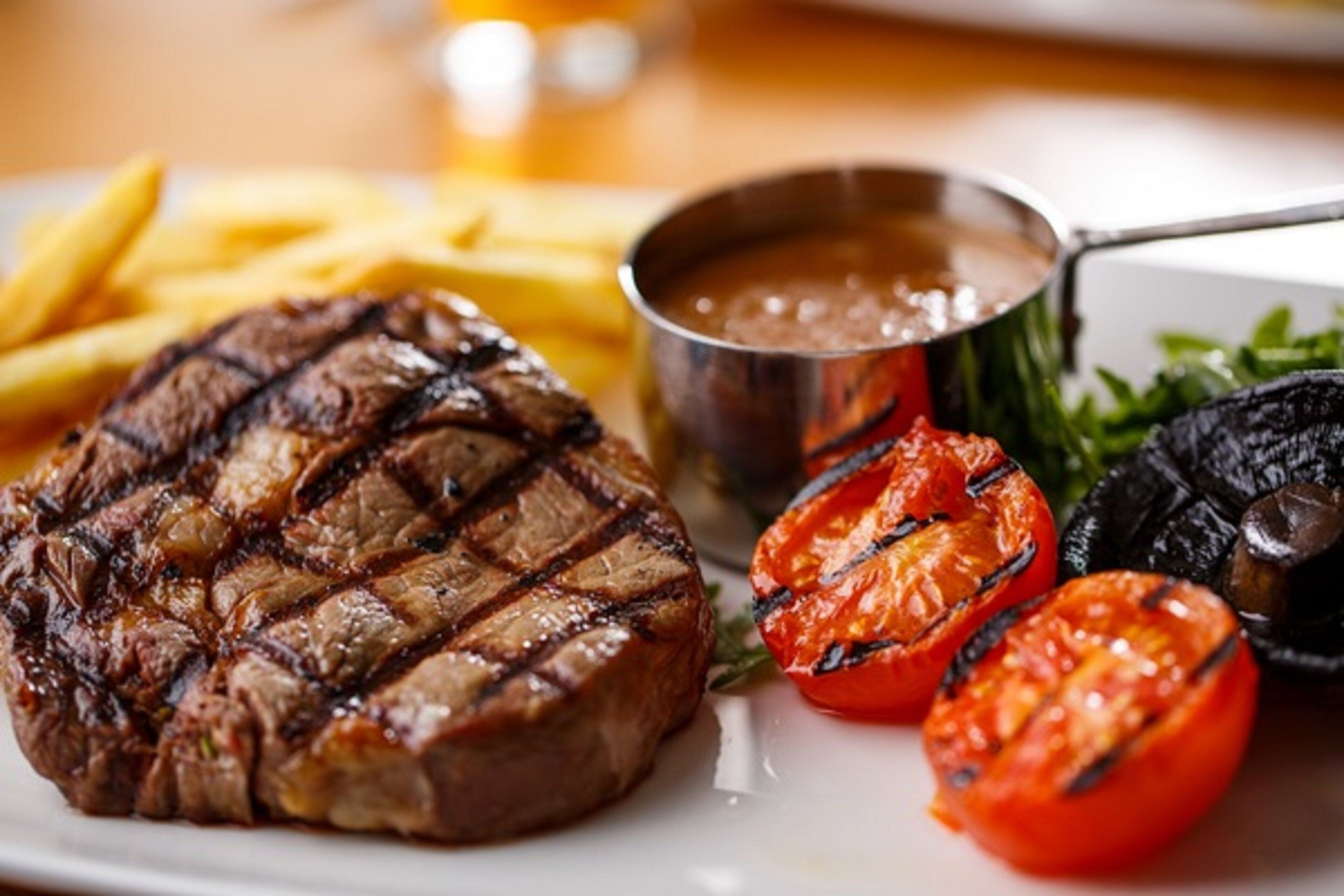 A steak with grill marks, French fries, grilled tomatoes, and a side of greens.