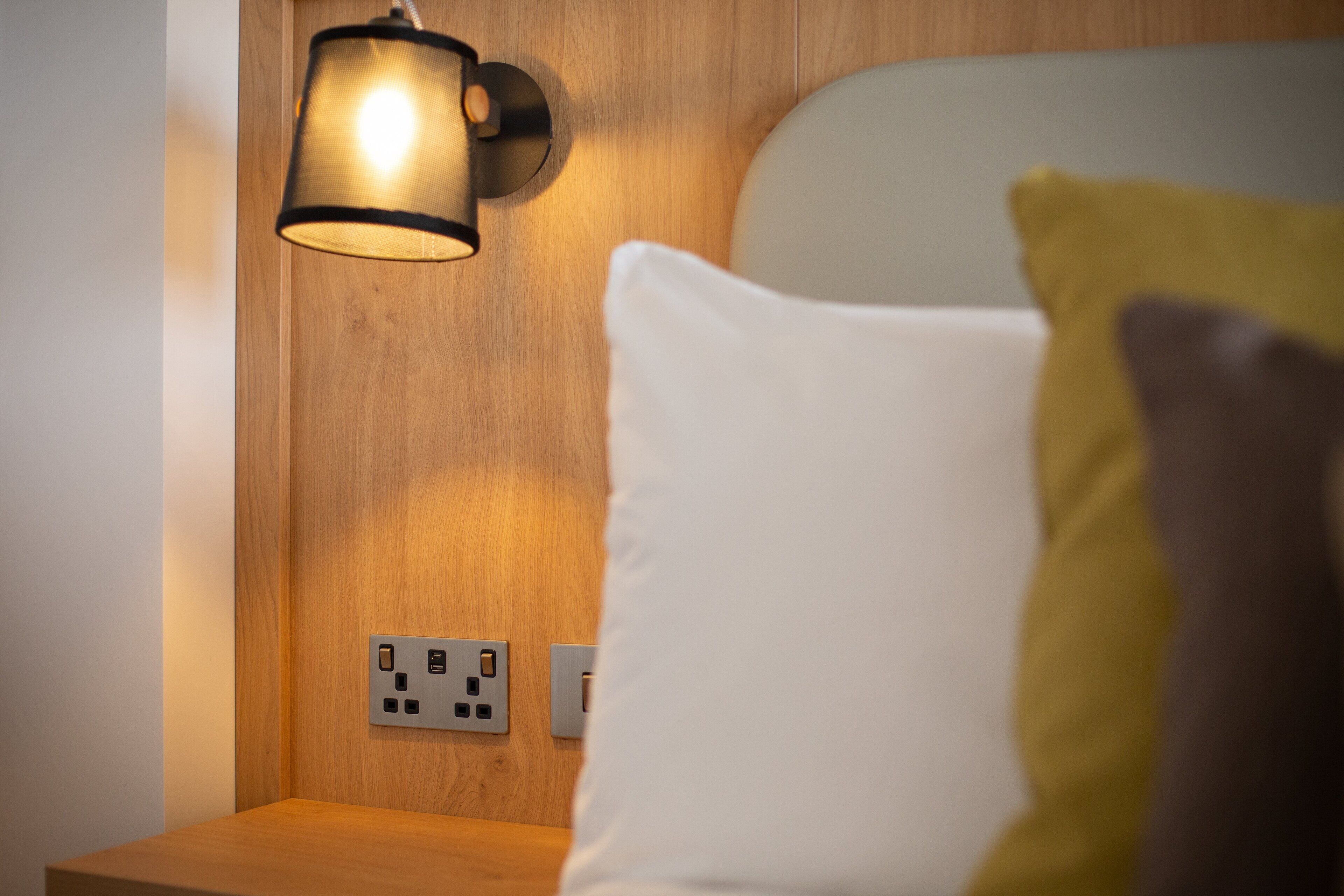 Een houten nachtkastje met een lamp, een stopcontact en een wit kussen.