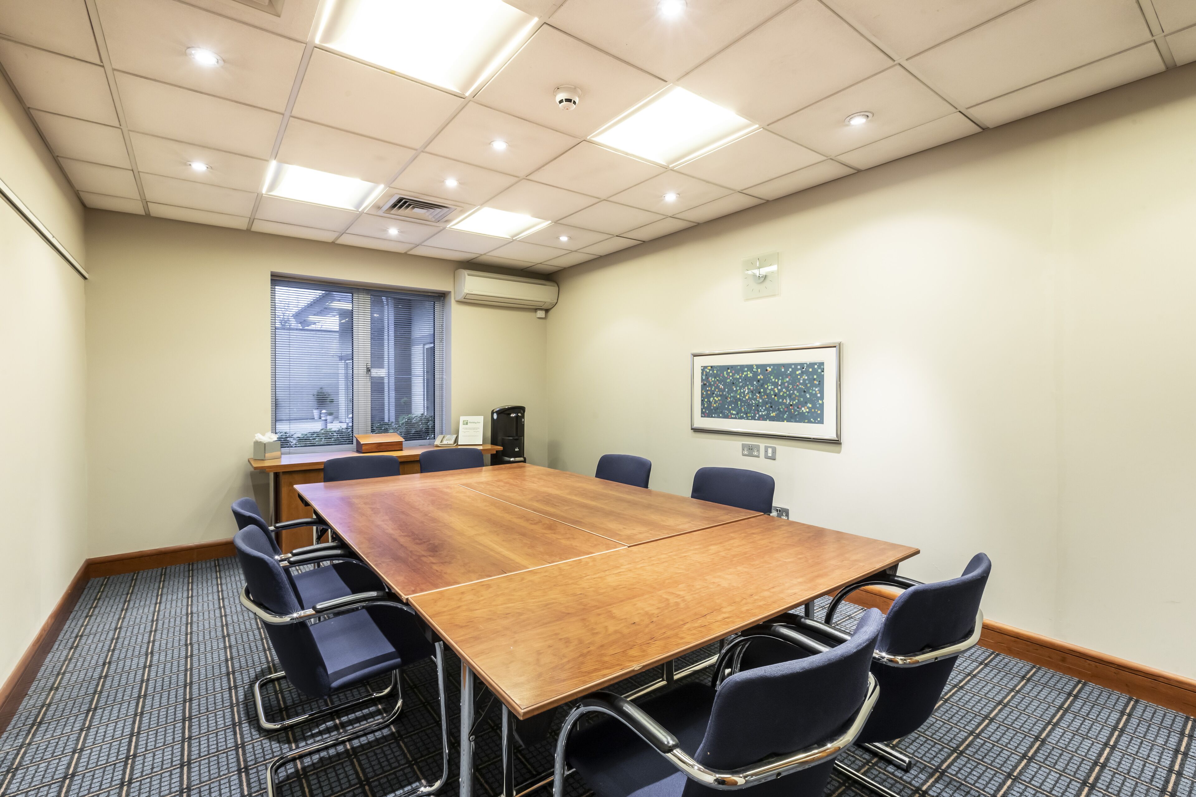 A conference room with a large wooden table, black chairs, a window with blinds, a framed picture on the wall, and an air conditioning unit.