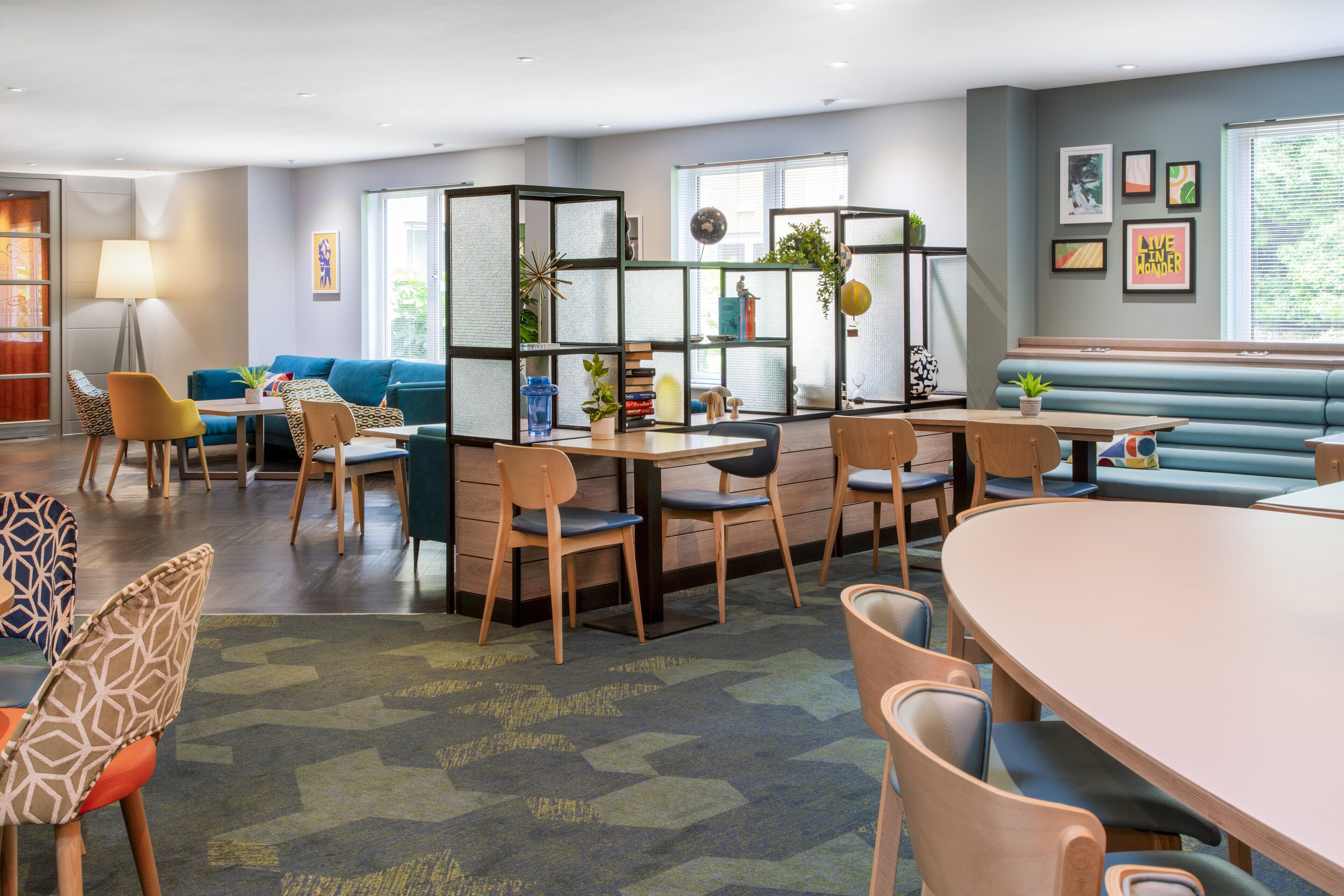 A modern lounge area with a round table, several chairs, a sofa, and a partition with plants and books.