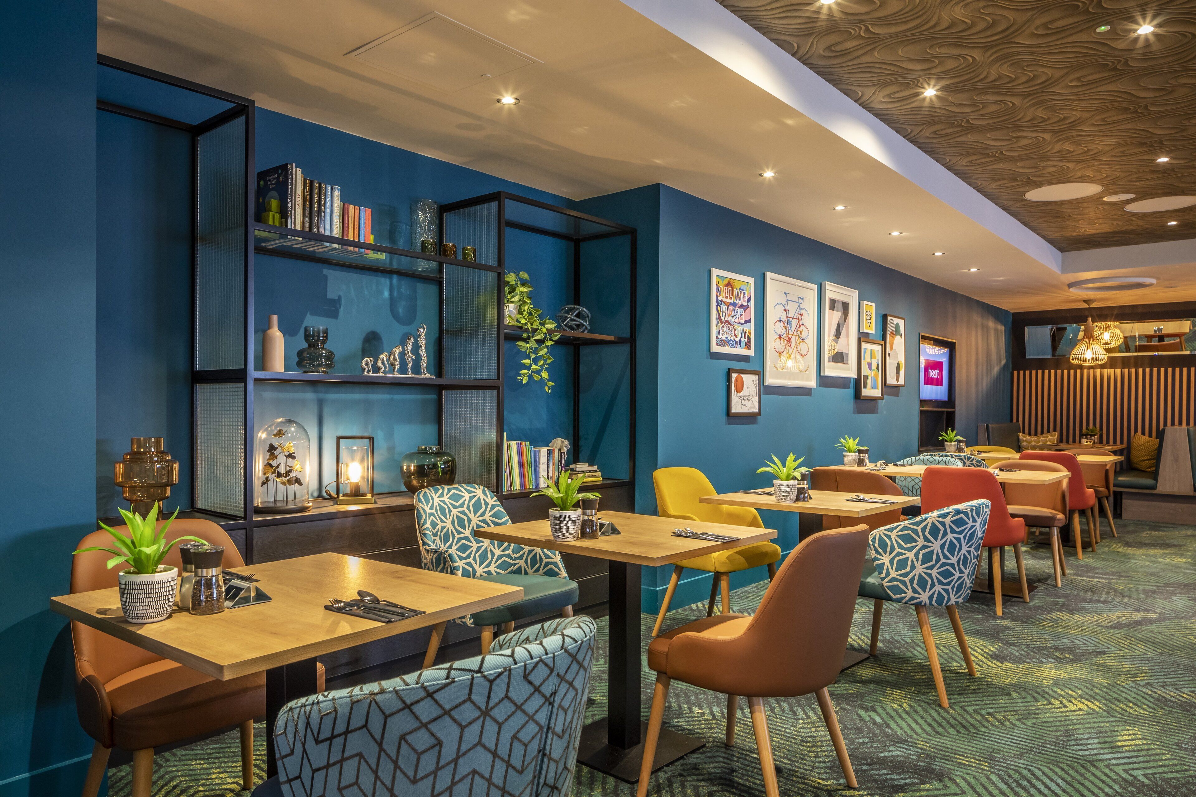 A modern dining area with tables and chairs, a bookshelf, and wall art.