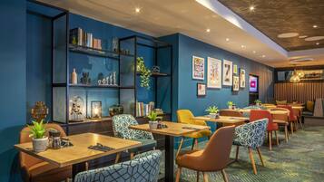 A modern dining area with tables and chairs, a bookshelf, and wall art.