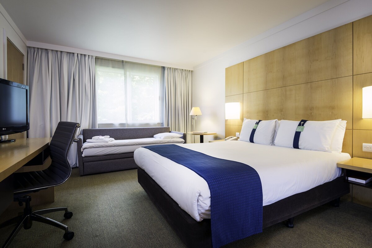 Una habitación de hotel con cama, escritorio, silla, televisión y ventana con cortinas.