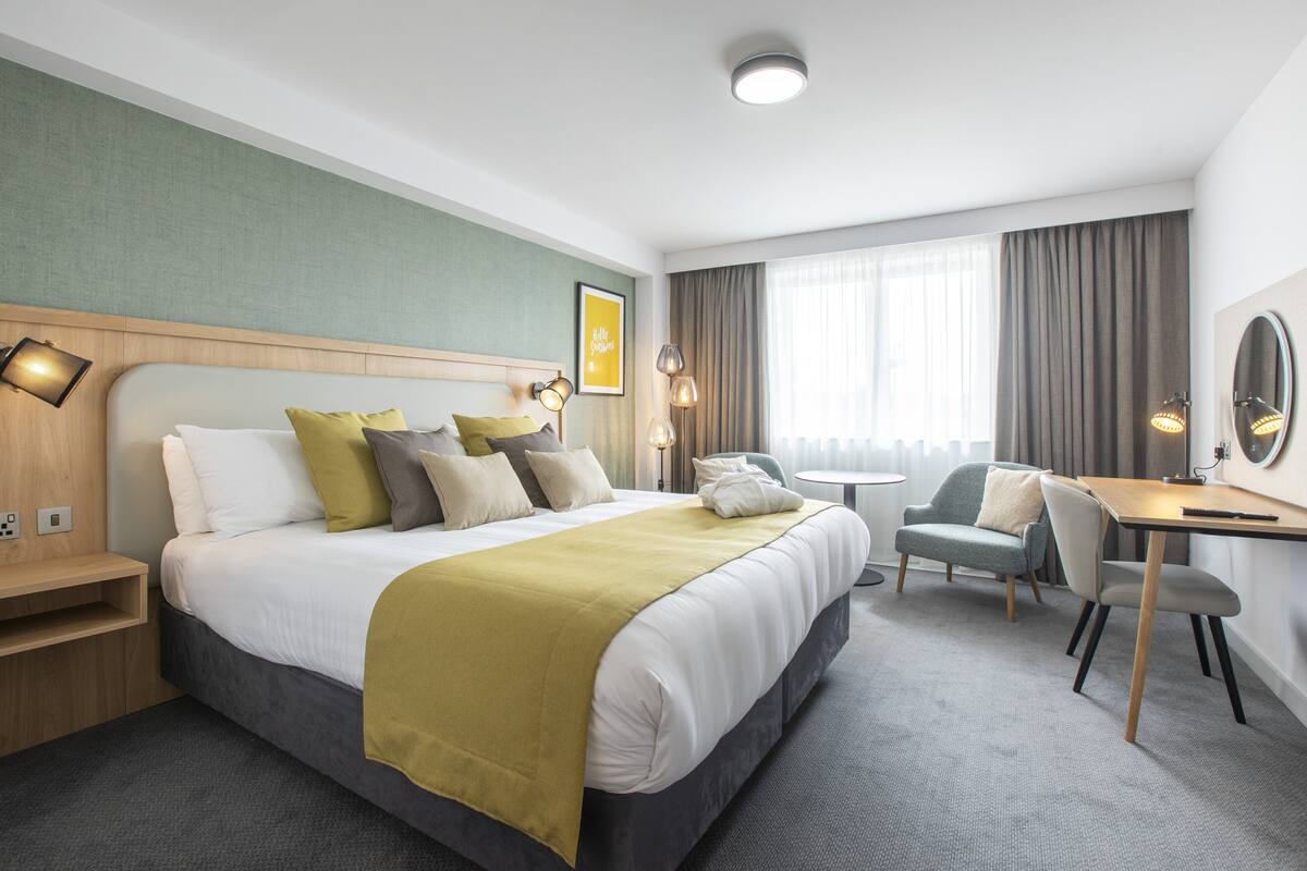 A hotel room with a large bed, a desk, and a chair. There is a window with curtains and a lamp on the desk.
