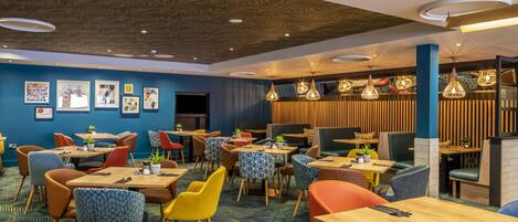 A modern restaurant with wooden tables and colorful chairs, blue walls, and hanging light fixtures.