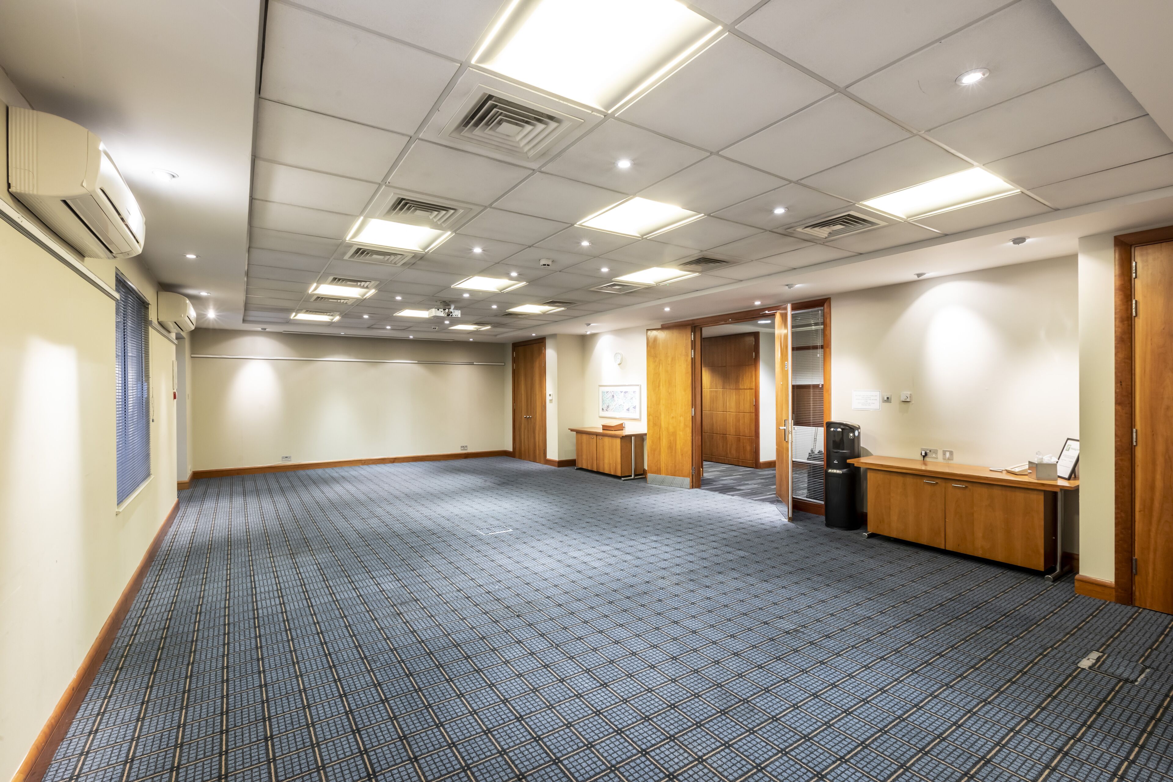 A large conference room with a carpeted floor, wooden paneling, and a kitchenette area.