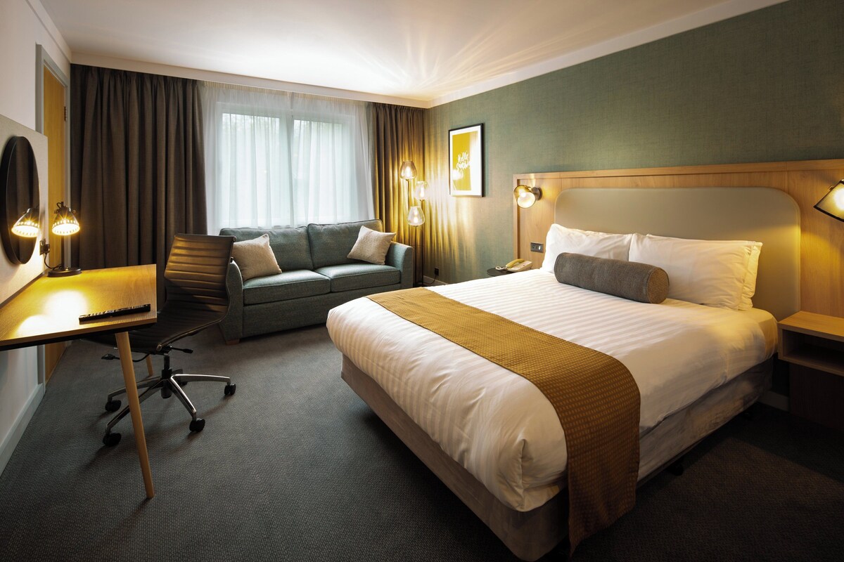 Habitación de hotel con una cama grande, un escritorio con lámpara, una silla, un sofá y una ventana con cortinas.