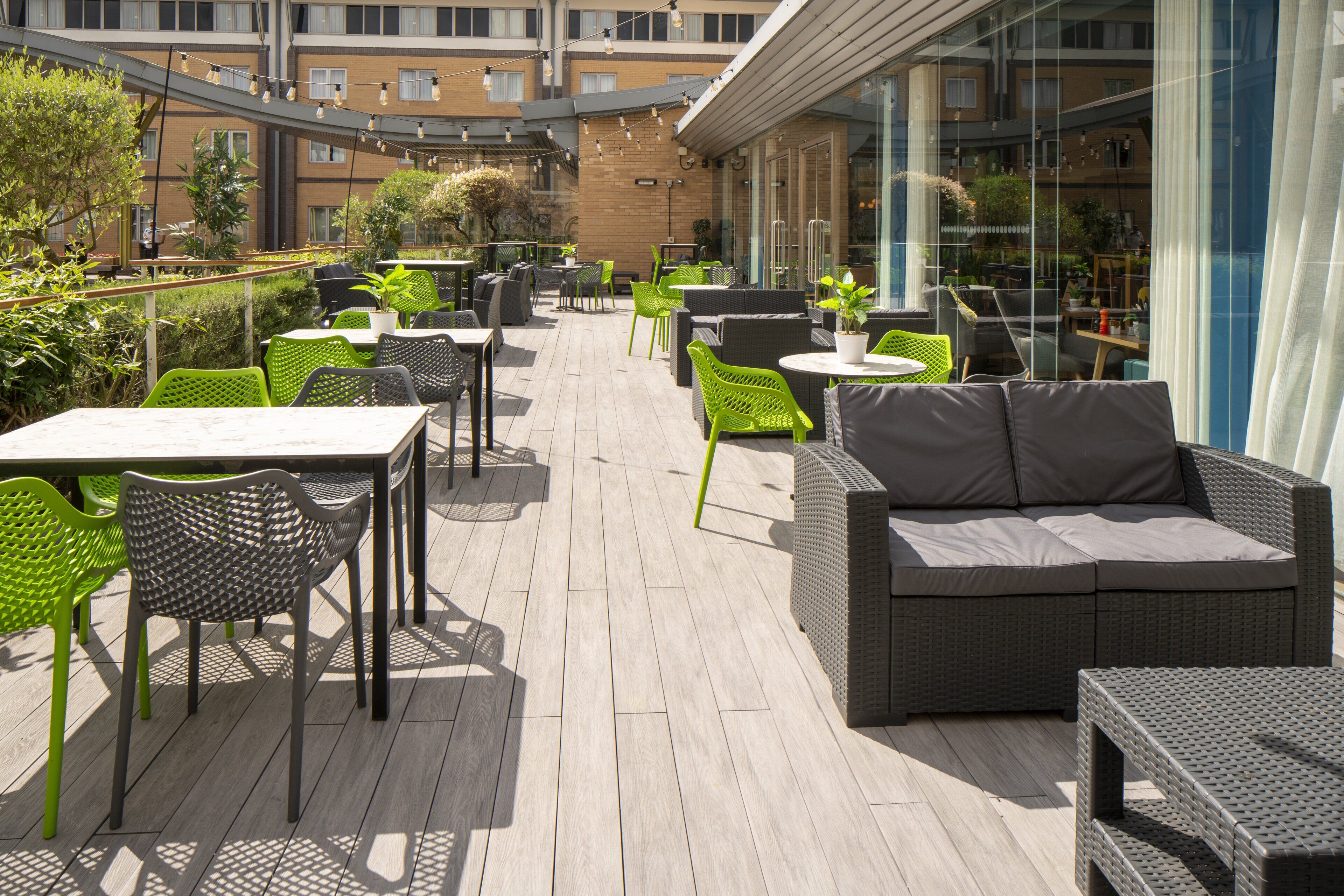 An outdoor patio with wicker furniture, tables, and chairs, surrounded by glass walls and a brick building in the background.