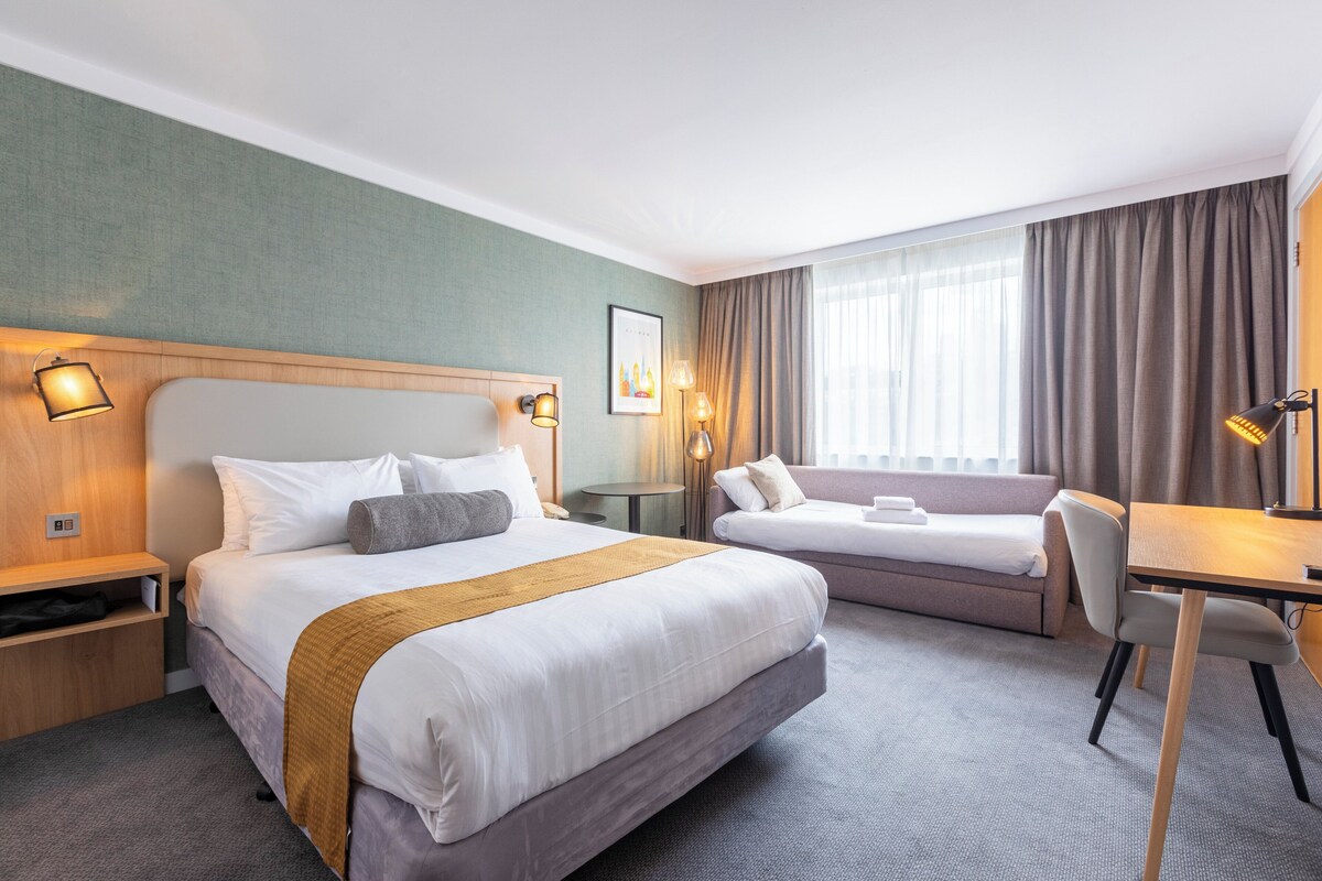 Habitación de hotel con una cama grande, otra más pequeña, un escritorio y una silla. Hay una ventana con cortinas, una lámpara y un cuadro en la pared.