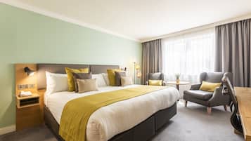 A hotel room with a large bed, bedside table, lamp, chair, and a small table with a plant.