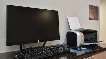 Un espace d’accueil d’hôtel comprenant un ordinateur de bureau, une imprimante et un panneau indiquant les équipements disponibles, tels que des toilettes, un distributeur de glaçons, un service de blanchisserie pour les clients et une salle Harison.