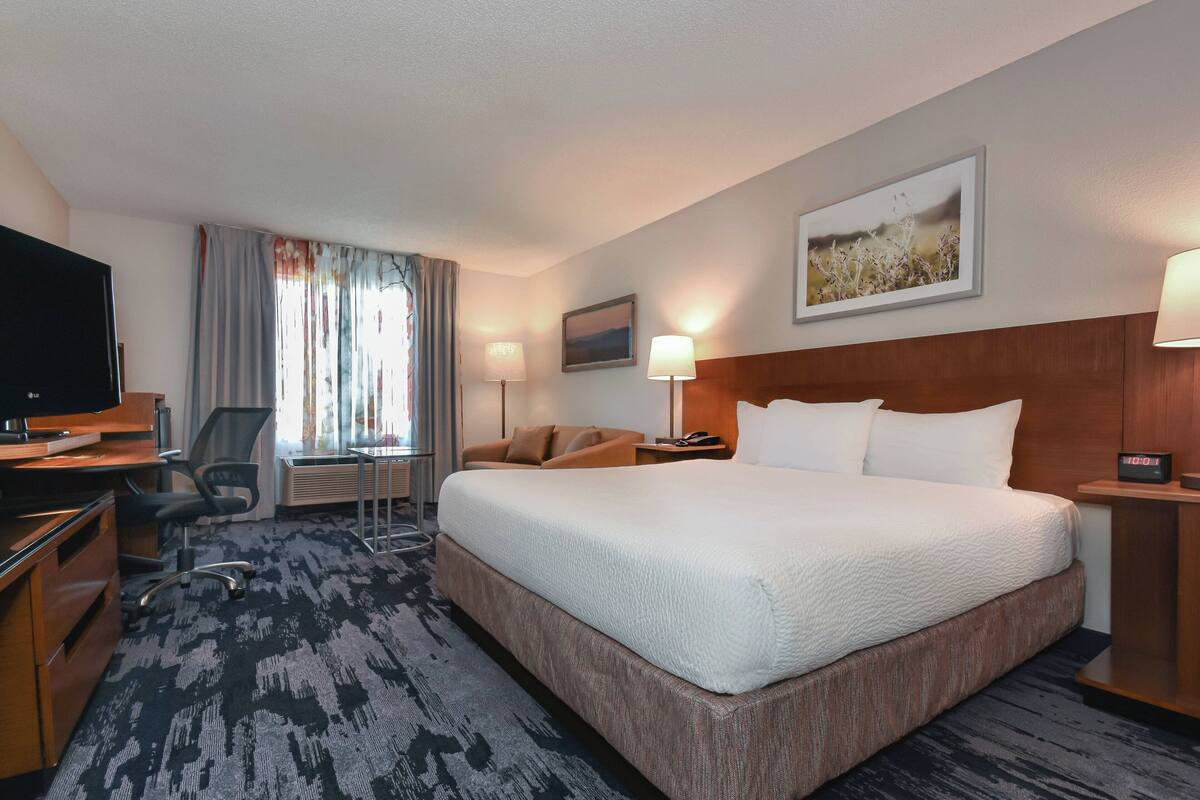 A hotel room with a large bed, a desk with a computer, a chair, a TV, and a window with curtains.
